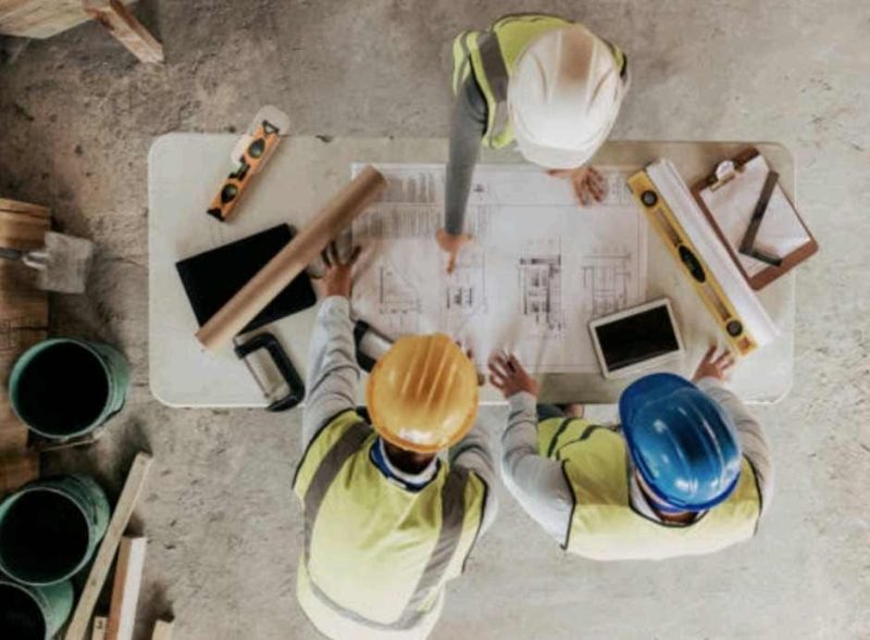 Ilustrasi pekerja konstruksi sedang memeriksa denah pembangunan rumah.
