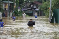 Sejumlah warga menyeberangi jalan yang terendam banjir tinggi saat hujan lebat melanda kawasan permukiman.