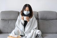 Seorang perempuan duduk di sofa dengan masker dan selimut sambil menikmati minuman hangat.