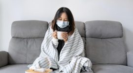 Seorang perempuan duduk di sofa dengan masker dan selimut sambil menikmati minuman hangat.