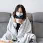 Seorang perempuan duduk di sofa dengan masker dan selimut sambil menikmati minuman hangat.
