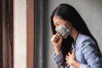 Perempuan tampak menahan batuk dan memegang dada, menunjukkan gejala gangguan pernapasan