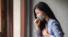 Perempuan tampak menahan batuk dan memegang dada, menunjukkan gejala gangguan pernapasan