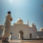 Seorang pria berdoa di halaman masjid megah dengan kubah putih dan menara tinggi di bawah langit cerah.