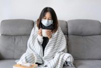 Seorang perempuan mengenakan masker dan selimut sambil memegang minuman hangat saat mengalami gejala flu di rumah.