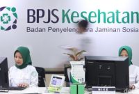 Petugas melayani peserta di kantor BPJS Kesehatan dengan latar logo BPJS Kesehatan.