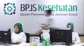 Petugas melayani peserta di kantor BPJS Kesehatan dengan latar logo BPJS Kesehatan.