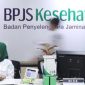 Petugas melayani peserta di kantor BPJS Kesehatan dengan latar logo BPJS Kesehatan.